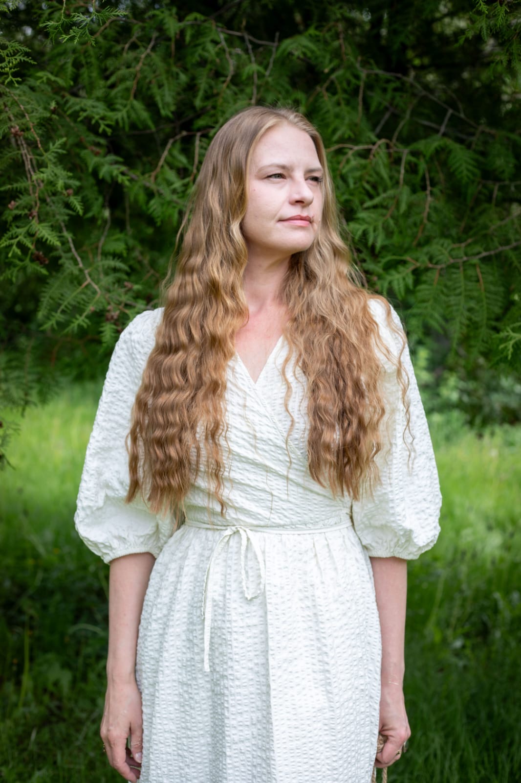 Woman with long wavy blonde hair wearing a white dress, standing outdoors before lush green foliage.
