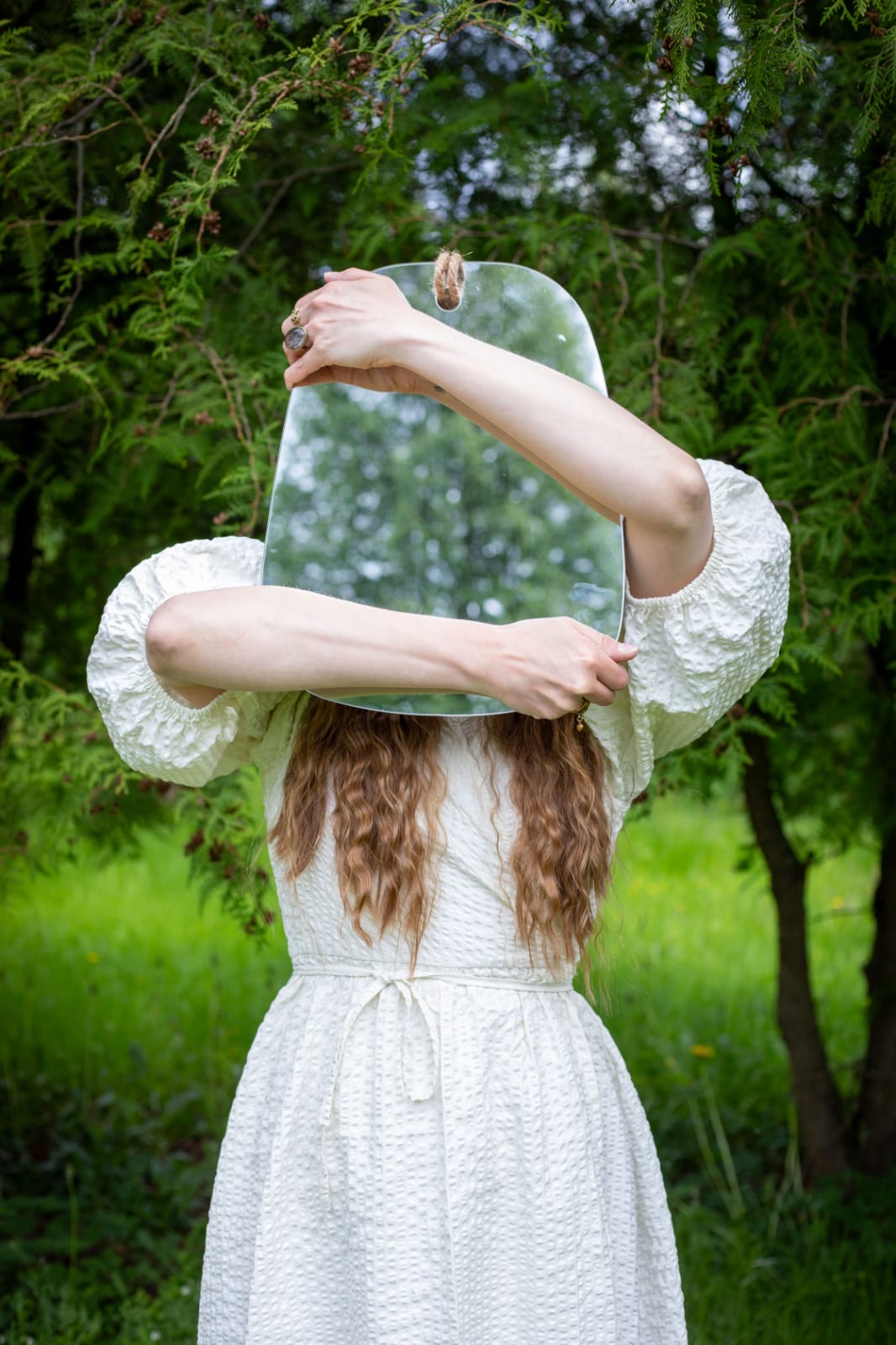 Woman in white dress holds a mirror on her face outdoors among lush green trees.