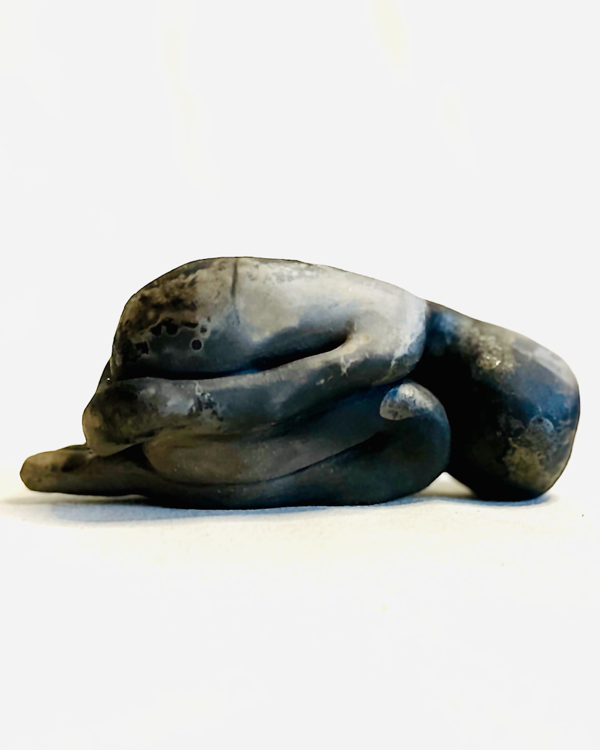Clay sculpture of a curled human-like character with dark and brown tones, resting on a white surface.