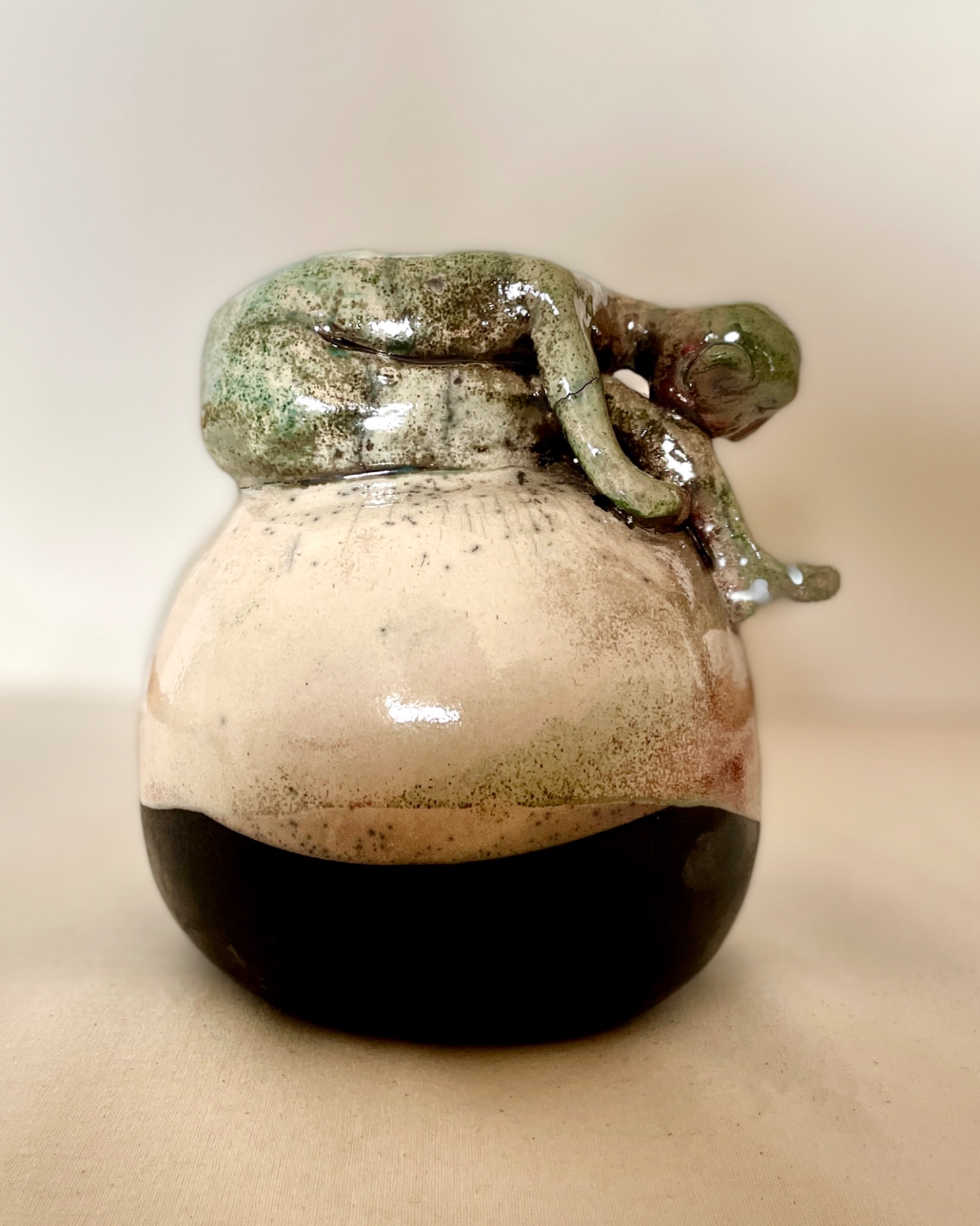 A raku-fired ceramic artwork against a light backround, featuring a spherical lower form with a human-like figure sitting on top, leaning against its legs. the piece is glazed but left unglazed near the bottom, with colors ranging from green to cream and accented with red details.