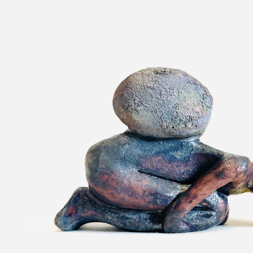 A raku-fired clay figure kneeling with a rock on its back. the image is cropped so that the figure's head is outside the frame.