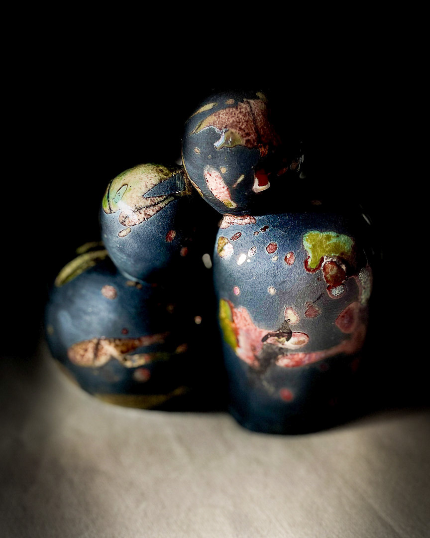 A ceramic artwork against dark background, featuring two round figures appearing to kiss. Thee piece has black surface with colorful splashes of glaze.