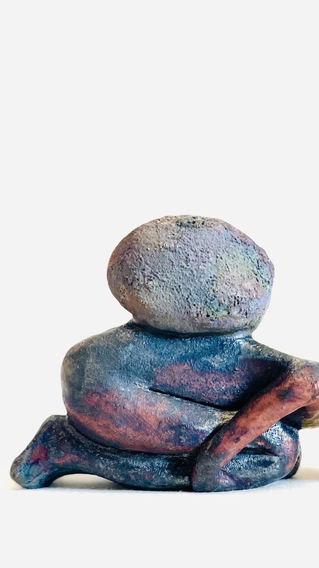 A raku-fired clay figure kneeling with a rock on its back. the image is cropped so that the figure's head is outside the frame.