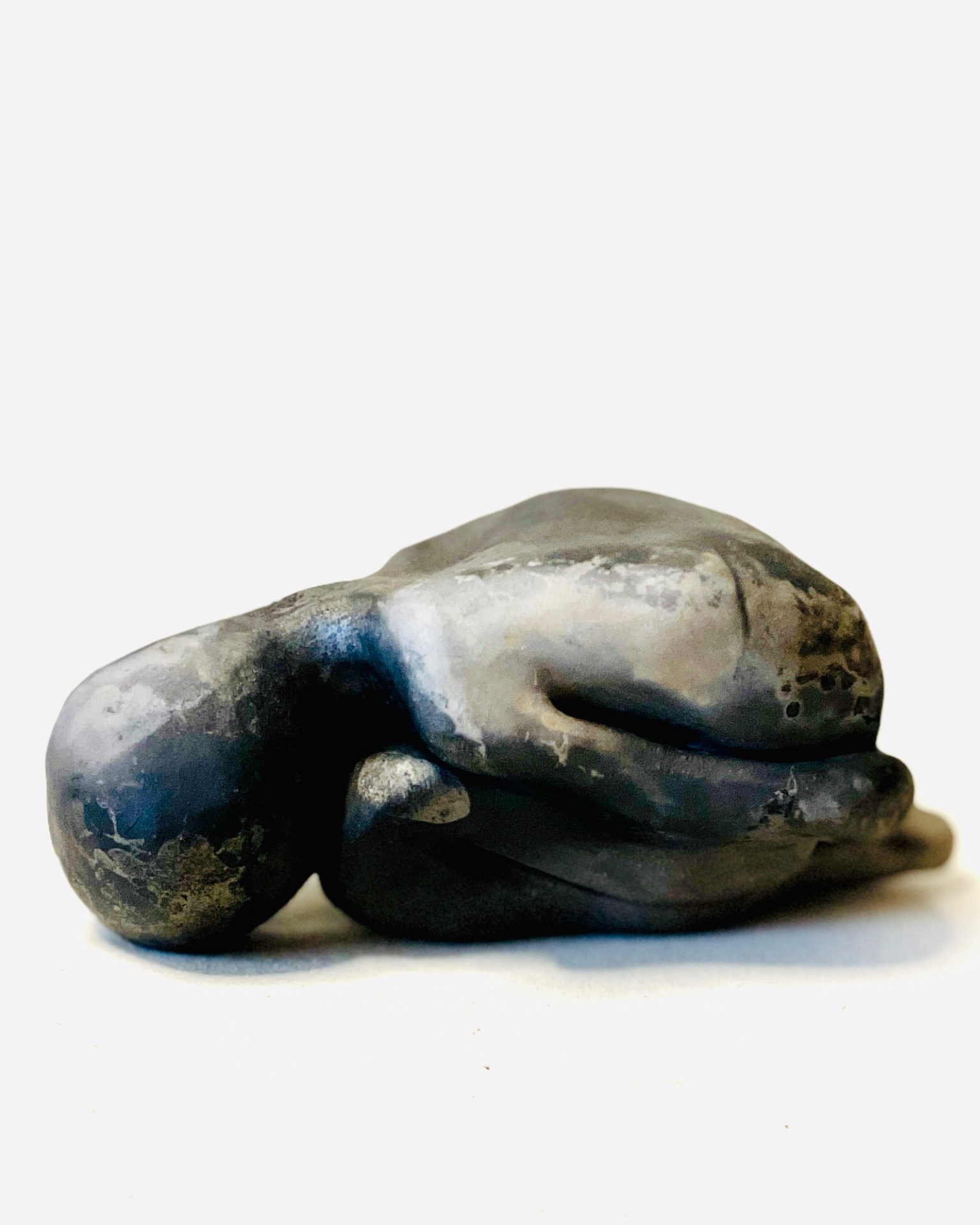 Clay sculpture of a curled human-like character with dark and brown tones, resting on a white surface.